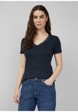 Kobieta o długich, falowanych brązowych włosach, ubrana w granatowy T-shirt z krótkim rękawem ozdobiony koronką oraz niebieskie dżinsy, stoi z jedną ręką w kieszeni.