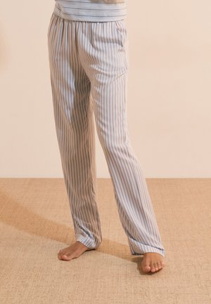 Persona con pantalones de pijama a rayas de color claro, de pie descalza sobre un suelo beige texturizado frente a una pared lisa.