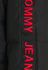 Tessuto nero con una striscia verticale nera testurizzata che presenta al centro un testo ricamato in rosso acceso con la scritta "Tommy Jean".