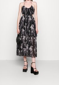 Nainen, joka on pukeutunut musta ja harmaa kukkakuosisella halterneck-mittapuvan, jossa on sivuleikkaukset, sekä korkea-alusteisiin kenkiin, ja hänellä on pieni musta käsilaukku.