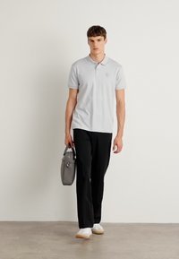 Camisola pólo cinza claro com mangas curtas e um logótipo discreto, combinada com calças pretas e sapatos brancos, segurando uma mala cinza.