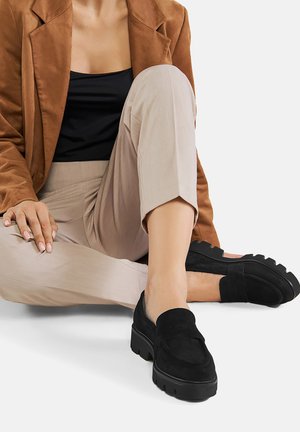 Schwarze Wildlederloafer mit dicker Plateausohle, runder Zehenform und genähten Details, getragen mit hellbeigen Hosen.