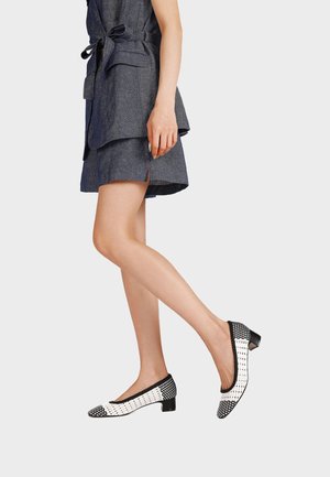 Donna che indossa un vestito grigio senza maniche con cintura e scarpe intrecciate bianche e nere con tacco basso, in piedi su uno sfondo semplice.