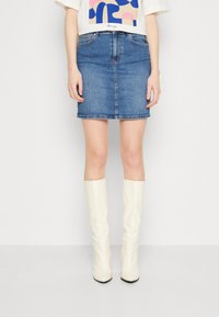 Denim minikjol i mellanblått med rak skärning, fem fickor och en fåll strax ovanför knäna, kombinerad med vita knähöga stövlar.