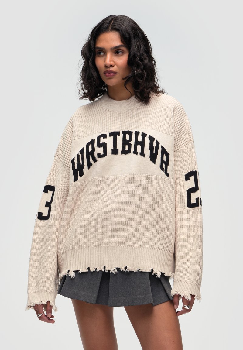 Beiger Strickpullover mit lockerer Passform, ausgestattet mit einem schwarzen Grafikdruck "WRSTBHVA" und "23" auf den Ärmeln, fransigem Saum und strukturiertem Stoff.