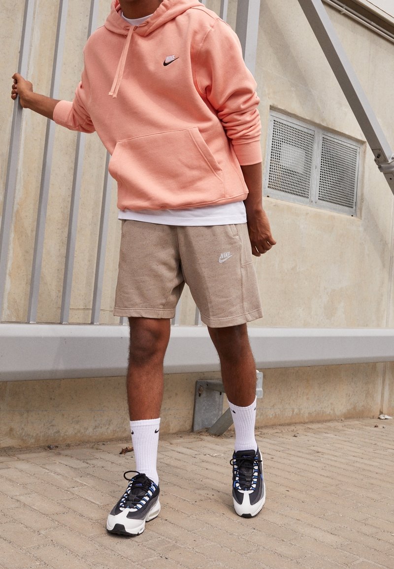 Hellrosa Nike Hoodie, beiges Shorts mit Logo, weiße Socken und schwarze Sneakers mit blauen Akzenten. Steht vor einem strukturierten Hintergrund.