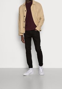 Beige linnedragt med sorte knapper, mørkerød T-shirt, sorte slim-fit bukser og hvide sneakers, præsenteret på en neutral baggrund.