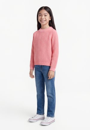 Smiling girl with long dark hair stands wearing a pink knit sweater, blue jeans, and white sneakers on a white background.