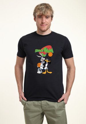 Junger Mann trägt ein schwarzes Space Jam T-Shirt mit Bugs Bunny und Daffy Duck, die Basketbälle halten, Hände in den Taschen, steht vor einfachem Hintergrund.