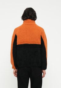 Chaqueta de forro polar con sección superior naranja y sección inferior negra. Tejido texturizado, cuello alto y corte holgado.