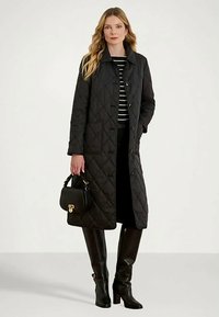 Frau in einem langen, schwarzen gesteppte Mantel, gestreiftem Hemd, schwarzen Hosen, hohen schwarzen Stiefeln und mit einer schwarzen Handtasche mit einem goldenen Verschluss.