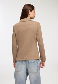 Giacca marrone, aderente, con colletto a pistagna, maniche lunghe e una texture liscia. Abbinata a jeans in denim azzurro chiaro, che mostrano uno stile casual.