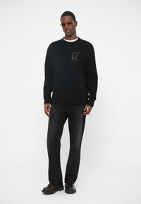 Pulover negru din tricot cu manșete și tiv cu țesătură în relief, având un logo pe piept. Asortat cu pantaloni din denim închis la culoare și pantofi negri.