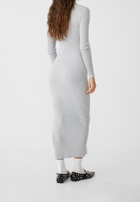 Abito in maglia grigia a coste con maniche lunghe, spacco laterale, design aderente, indossato con scarpe nere con borchie decorative e calze bianche.