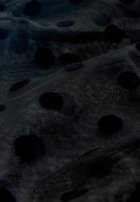 Sort gennemsigtigt stof med et tekstureret netdesign, der har forhøjede sorte prikker i forskellige størrelser, der er arrangeret sporadisk over overfladen.