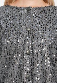 Zilveren kledingstuk versierd met pailletten en een gestructureerd oppervlak; heeft twee donkere knopen en een ronde halslijn. Sprankelend, reflecterend materiaal.