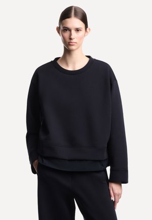 Giovane donna con capelli scuri raccolti in uno chignon basso, indossa una felpa nera larga e pantaloni neri, in piedi davanti a uno sfondo bianco semplice.