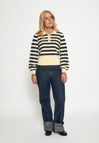 Prúžkovaný pulóver v námorníckej modrej a žltej farbe, s golierom a gombíkmi, kombinovaný s tmavými riflami, mierne voľný s prehnutými manžetami.