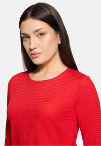 Czerwony top z długim rękawem wykonany z teksturowanej tkaniny, z okrągłym dekoltem. Intensywny kolor z wyraźnym, prążkowanym wzorem na całej powierzchni.