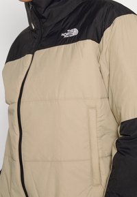 Puffjacka med en beige kropp och svarta axlar. Har en framzipper, quiltrad textur och The North Face-logotyp på bröstet.