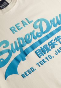 Camiseta de algodón crema con texto gráfico en relieve de color azul, "REAL SuperDry" y caracteres japoneses, con textura suave y cuello redondo.