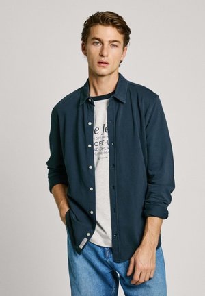 Chemise boutonnée bleu marine en mélange de coton, avec un col ouvert, des poches frontales et une texture douce, portée par-dessus un t-shirt graphique gris.