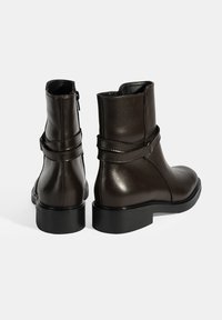Bottines en cuir marron avec une texture lisse, dotées d'une fermeture éclair sur le côté, d'un simple détail de boucle et d'une semelle plate noire.