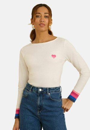 Langärmeliges cremefarbenes Hemd mit einer rosa Herzgrafik, kombiniert mit hoch taillierten blauen Jeans aus Denim. Die Ärmelbündchen zeigen kräftige Streifen in Rosa und Blau.