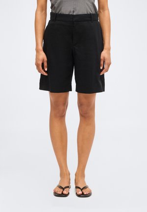 Osoba w czarnych szortach do kolan i czarnych sandałach, stojąca na jednolitym jasnym tle.