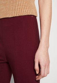 Stickad tan sweater med ribbad textur, bärs över åtsittande burgundy-byxor. Synlig hand som vilar mot byxornas släta yta.