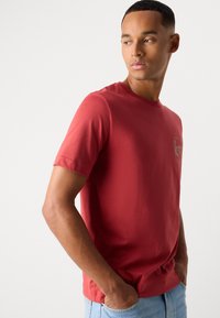 Moški v rdeči majici s kratkimi rokavi in svetlo modrih jeansih, stoji z rokama v žepih in gleda desno proti enotni ozadju.