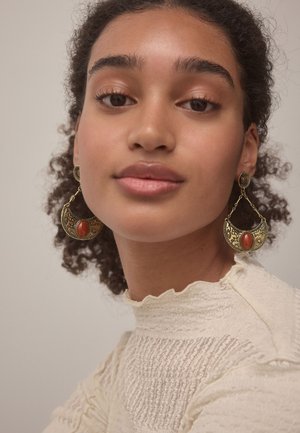 Young woman with curly hair wears ornate gold and red crescent-shaped earrings and a cream textured high-neck top.