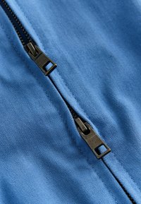 Gros plan d'une fermeture éclair métallique noire partiellement ouverte sur un tissu bleu lisse, montrant les dents et les curseurs de la fermeture éclair.