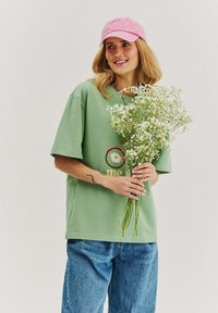 Woman in green shirt and pink cap holding a bouquet of small white flowers, smiling and looking slightly to the side.