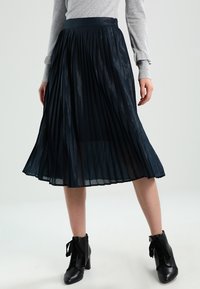 Námořnická plisovaná sukně s lesklou texturou, vysokým pasem a volným designem, doplněná černými kotníkovými botami s mašlemi.