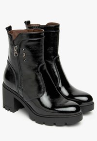 Bottines noires en matériau brillant, avec une fermeture éclair latérale, un talon épais et une semelle en caoutchouc texturée. Les accents incluent des clous métalliques.
