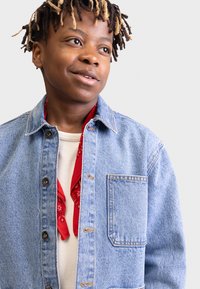 Denim jacket in light blue with a front pocket, button closure, and contrasting red bandana peeking out from underneath a cream shirt.