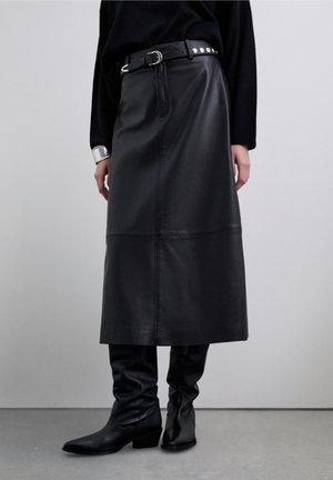 Falda midi de cuero negro con una silueta recta, que presenta una textura lisa y un cinturón con tachuelas plateadas. Combinada con botas negras hasta la rodilla.