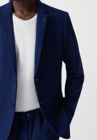 Granatowa, strukturalna marynarka z wyłogami, dwoma guzikami i jednym ozdobnym guzikiem. Połączona z gładką białą koszulą i dopasowanymi spodniami.