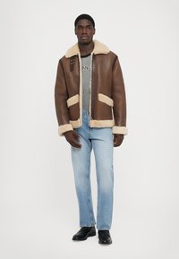 Veste en shearling marron avec des finitions crème, fermeture éclair à l'avant, deux poches plaquées, portée sur un t-shirt gris et un jean bleu clair, accompagnée de chaussures noires.