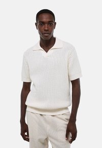 Chemise polo blanche à manches courtes avec texture côtelée et col en V, associée à un pantalon texturé de couleur claire.