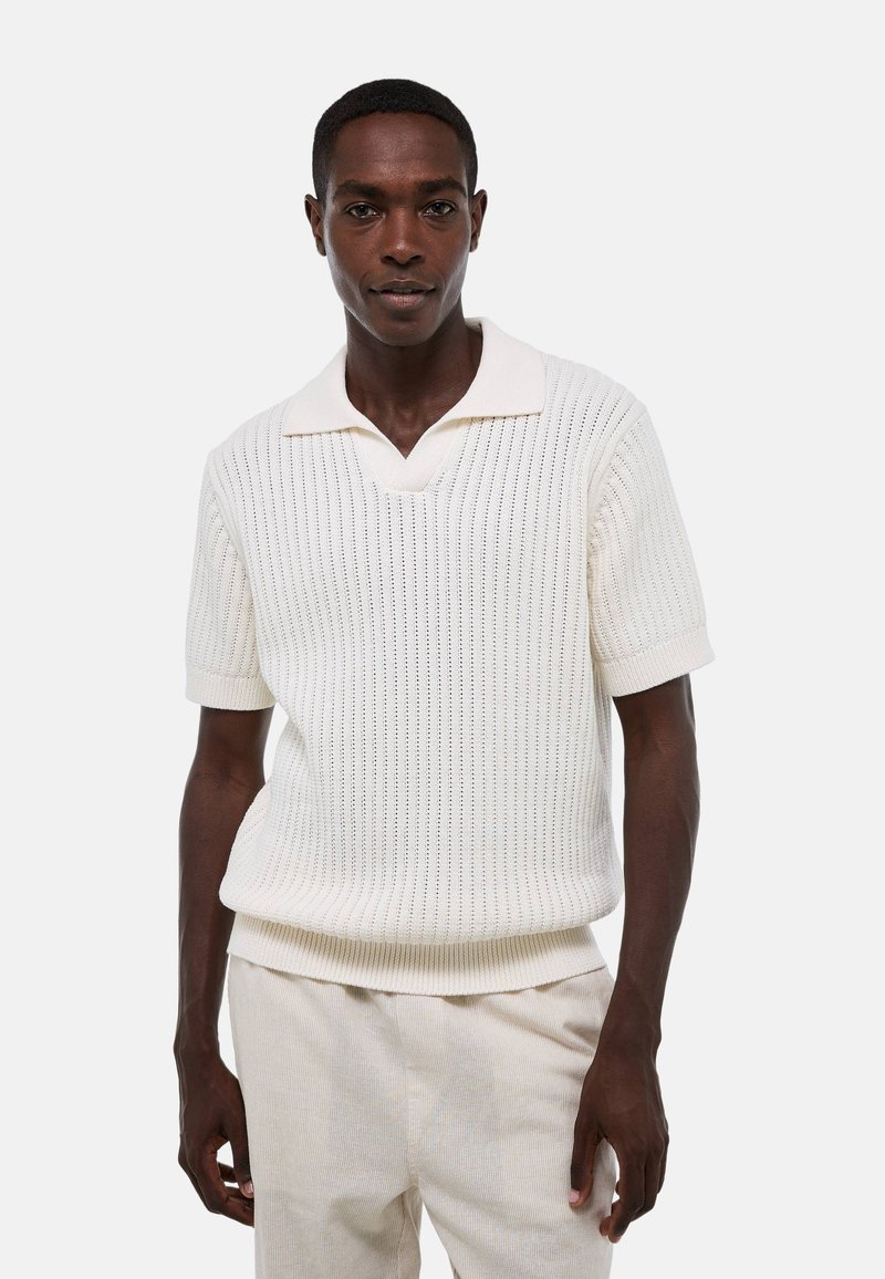 Chemise polo blanche à manches courtes avec texture côtelée et col en V, associée à un pantalon texturé de couleur claire.