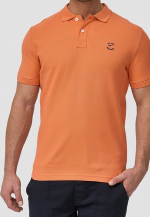 Mężczyzna w pomarańczowej koszulce polo z krótkim rękawem, z dwoma guzikami i małym granatowym logo na lewej piersi, w zestawieniu z ciemnymi spodniami i jedną ręką w kieszeni.