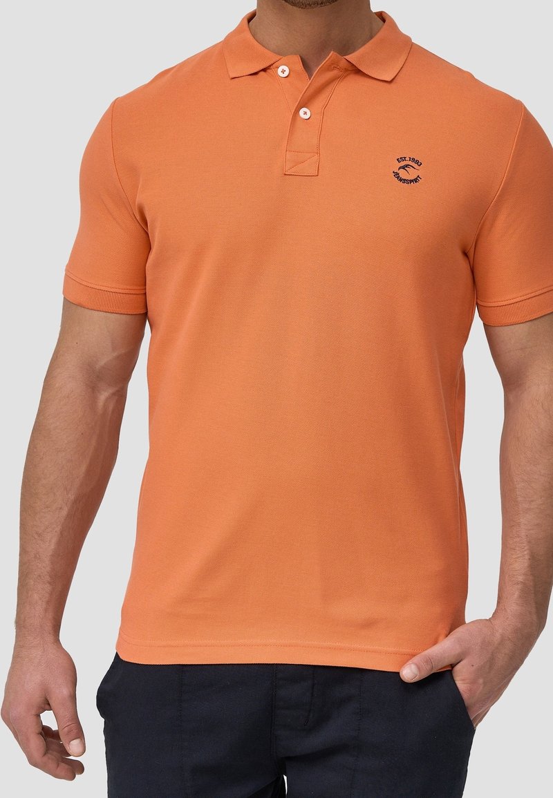 Homme portant un polo orange à manches courtes avec deux boutons et un petit logo bleu marine sur la poitrine gauche, associé à un pantalon foncé et une main dans la poche.