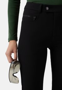 Black pants with a zippered pocket, paired with a green long-sleeve top. A pair of sunglasses is held in the right hand.