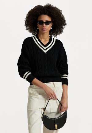 Black cable-knit sweater with white striped V-neck and cuffs, paired with cream-colored pants and a curved black handbag. Model wearing sunglasses.