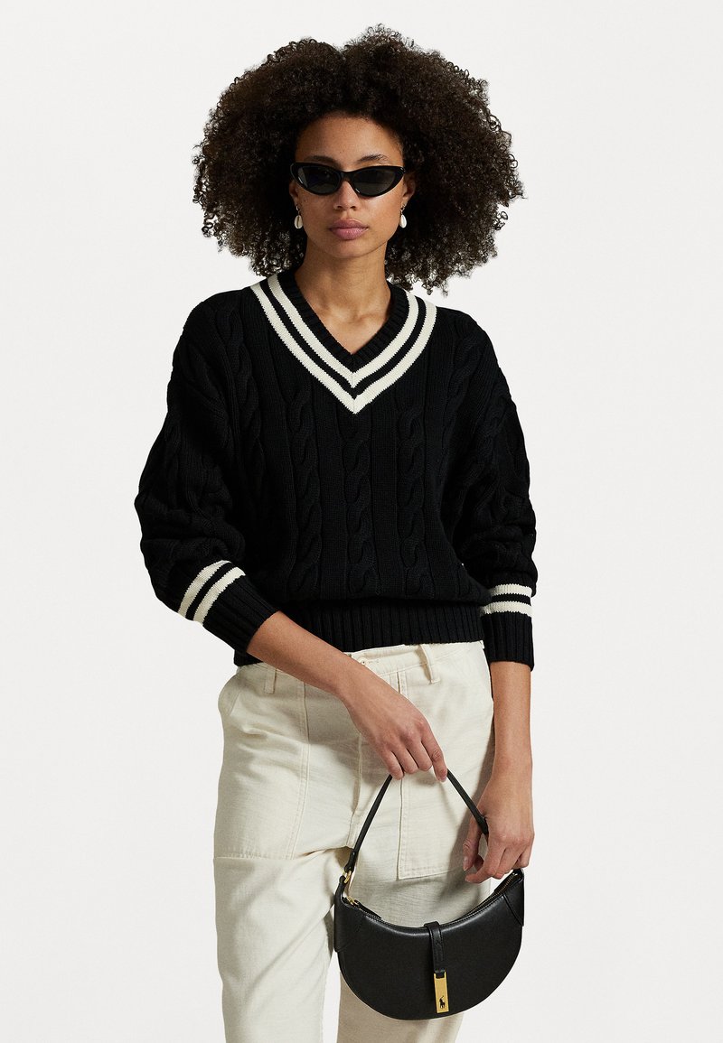 Pull en tricot noir avec un col en V rayé blanc et des poignets, associé à un pantalon crème et un sac à main noir arrondi. Modèle portant des lunettes de soleil.