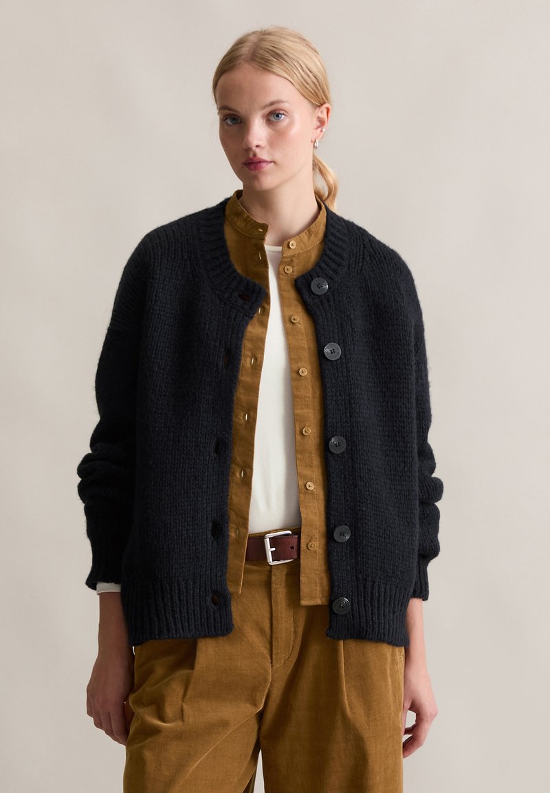 Cardigan nero a maglia con scollo rotondo e bottoni, abbinato a una camicia gialla con bottoni e pantaloni marroni. Texture liscia e vestibilità comoda.