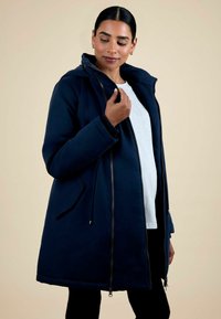 Cappotto con cappuccio blu navy in tessuto morbido, con chiusura a zip frontale, tasche laterali e coulisse regolabile in vita, indossato sopra una maglietta grigia chiara.
