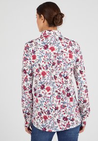Donna con un'acconciatura a bun castano, che indossa una camicia bianca a maniche lunghe con fiori e jeans blu, vista di spalle.